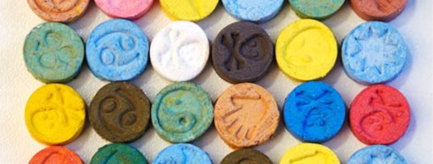 A variety of colorful MDMA (Molly) tablets arranged on a white surface, representing the different forms in which the drug is consumed.