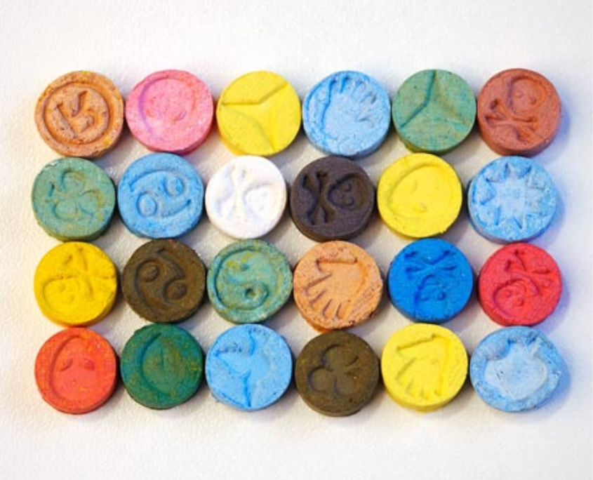 A variety of colorful MDMA (Molly) tablets arranged on a white surface, representing the different forms in which the drug is consumed.