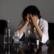 A lesbian person looking confused and anxious sitting on a table with scattered white pills and water.