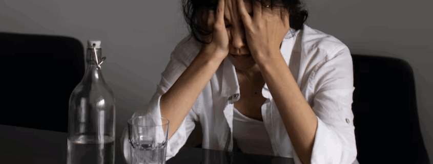 A lesbian person looking confused and anxious sitting on a table with scattered white pills and water.